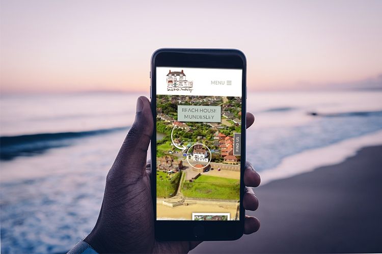 A hand holding a mobile phone is displayed on a beach at dusk. The Beach House Mundesley website is shown on the mobile phone and draws your attention to the centre of the screen.