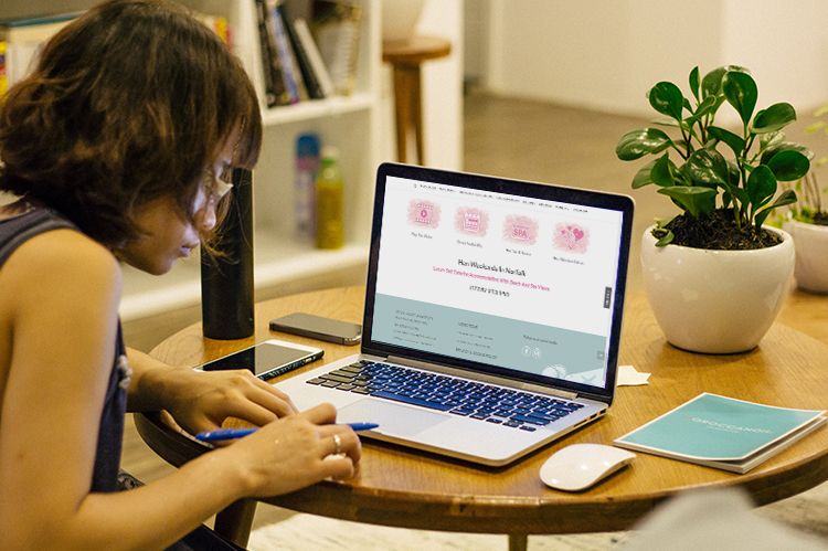 A woman is making notes sitting at a table. On the table lies a laptop displaying the Beach House Mundesley website. There is a potted pant on the table.