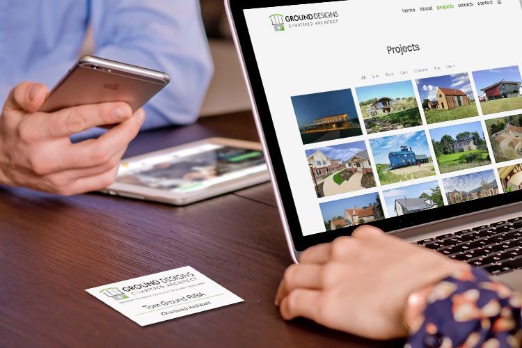 A man and woman look at electronic devices. The laptop displays the Ground Designs Chartered Architect website. There is a business card on the table.