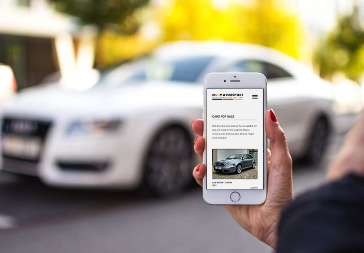 Woman holding a mobile phone showing a MCP Motorsport car listing page. A car can be seen in the background.