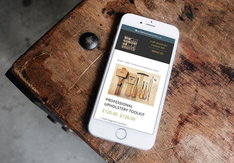 A mobile phone lies on an old wooden piece of furniture and displays the School of Stuff website.