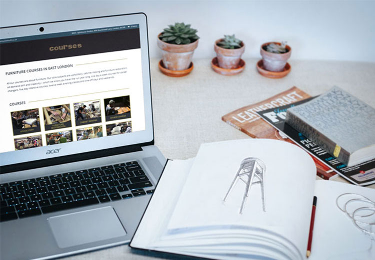 A laptop displaying the School of Stuff website sits on a desk next to an open book with hand drawn sketches of a bar stool.