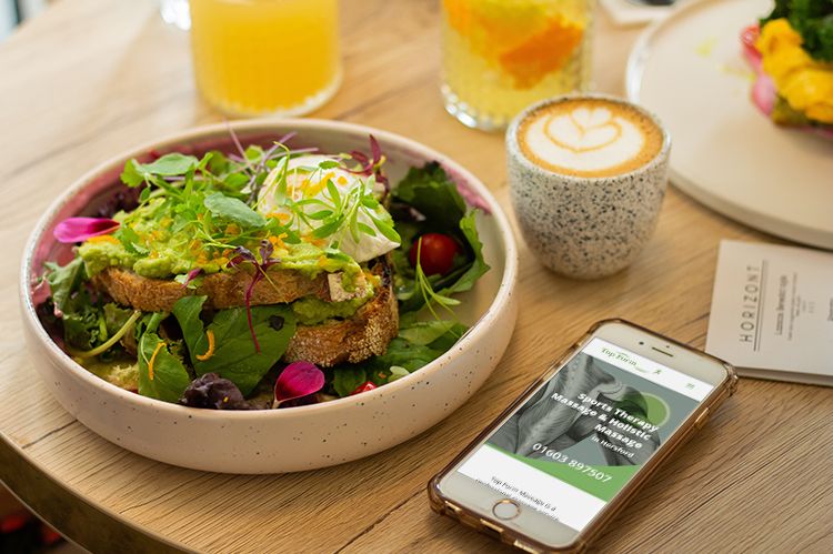 The Top Form Massage website displayed on a mobile phone, which lies on a dining table, with a healthy salad in a bowl and a cup of coffee,