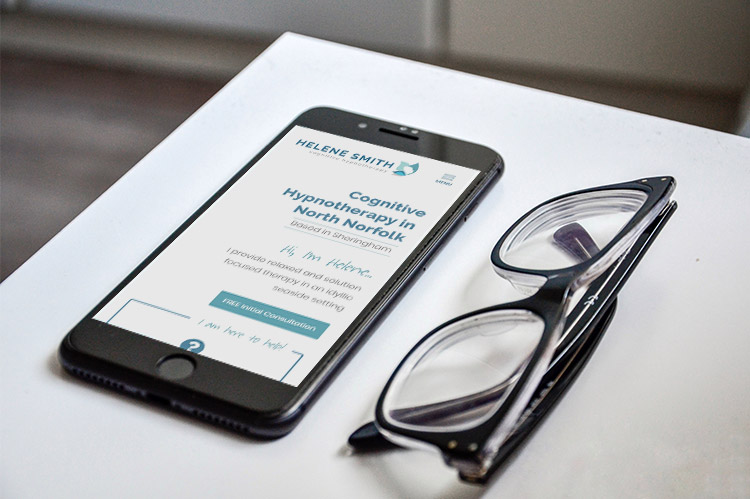 A mobile phone showing the Helene Smith Cognitive Hypnotherapist website, next to a pair of reading glasses on a desk.