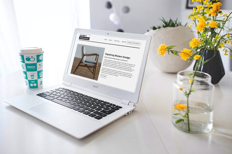 A laptop on a table showing the Modern Restoration website. Beside the laptop there is some yellow flowers in a glass jar and paper coffee cup.