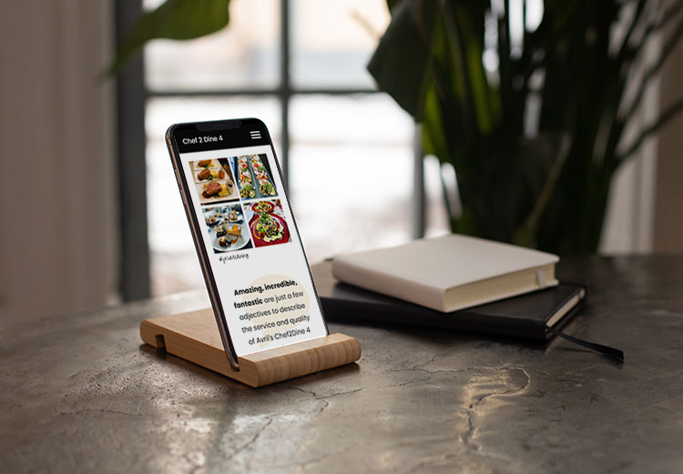A mobile phone displaying a Chef 2 Dine 4 webpage on a kitchen worktop stand.