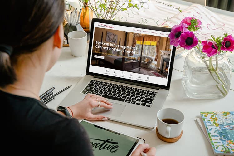 Woman looking at a laptop on her desk viewing a holiday home website. A cup of coffee and some pink flowers are on the table.